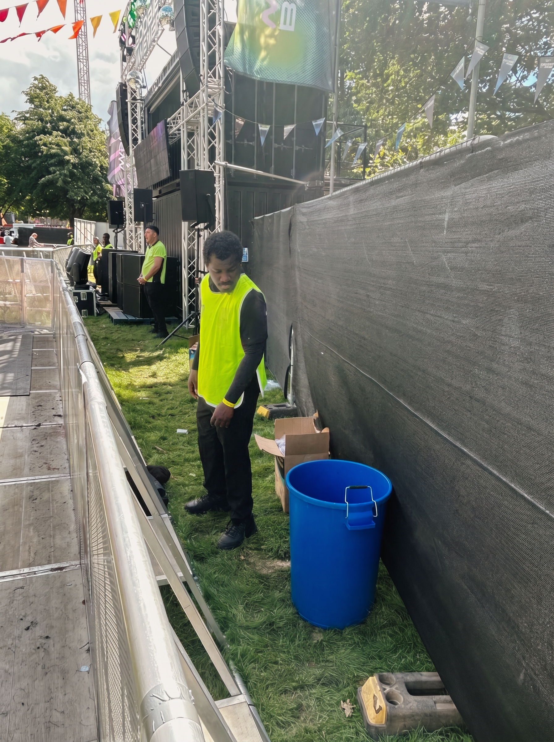 Steward standing beside an open blue bin and a cardboard box of cups at the pit barrier